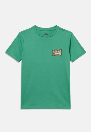 Grøn T-shirt med korte ærmer og rund hals, med en lille, mønstret lap på venstre bryst, der har et stiliseret logo og tekst.