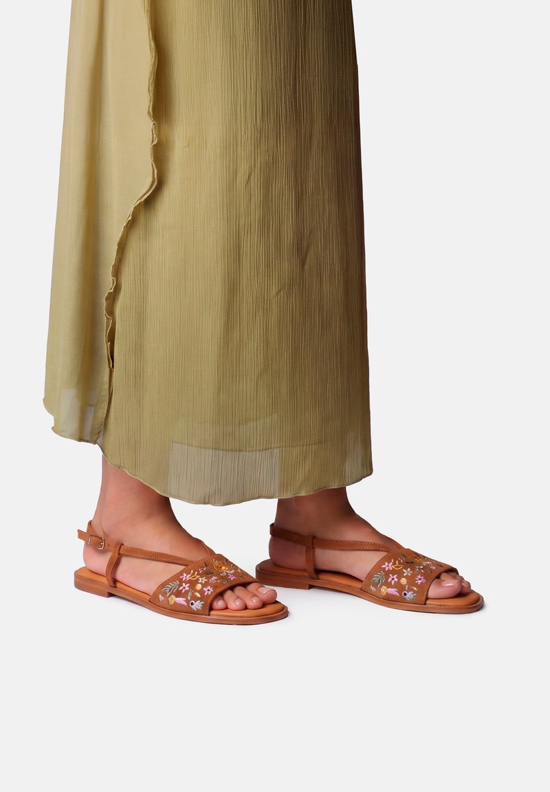Pieds portant des sandales en cuir marron avec des fleurs brodées et des brides à la cheville, visibles sous une longue jupe vert olive clair.