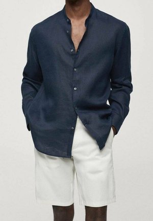 Man wearing a navy blue mandarin-collar long-sleeve shirt with white knee-length shorts, hands in pockets, against a plain light background.