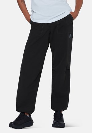 Person trägt schwarze, lockere Hose mit elastischen Bündchen und schwarze Sneakers, Hände in den Taschen, stehend vor weißem Hintergrund.
