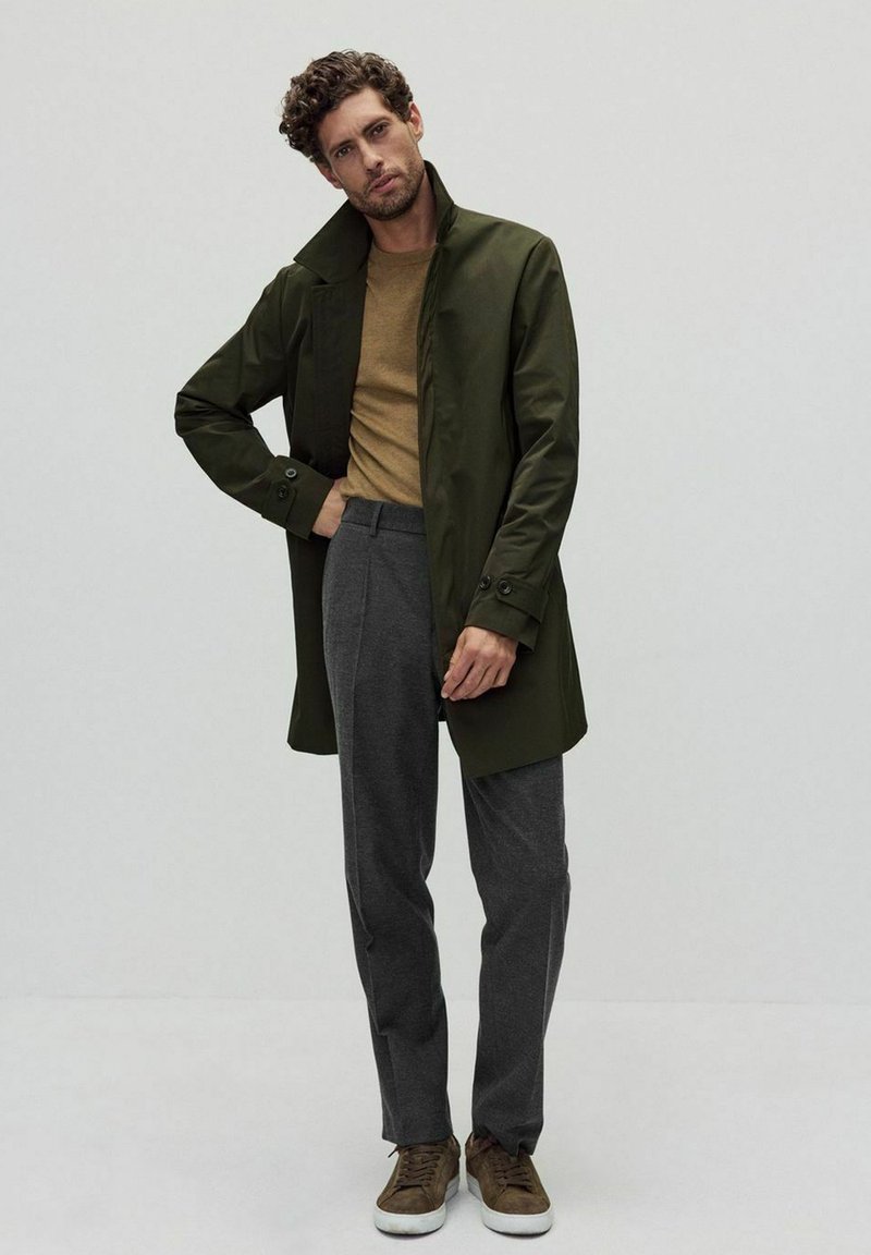 Vihreä vedenpitävä takki, jossa on korkea kaulus, ruskea neule sen alla, harmaat räätälöidyt housut ja ruskeat kengät. Sujuva materiaali, jossa on rakenteellinen istuvuus.