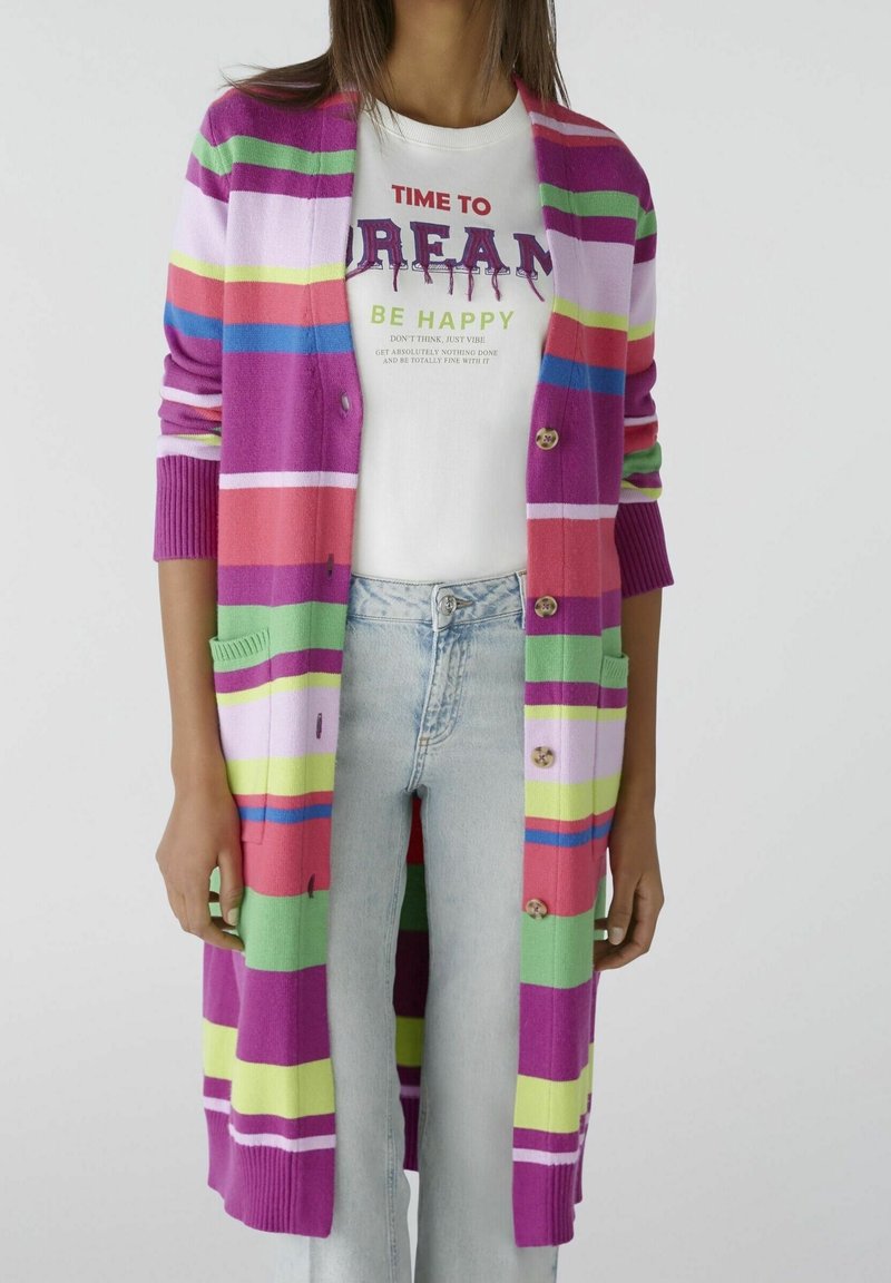 Femme portant un long cardigan rayé rose, vert, violet et bleu par-dessus un t-shirt blanc avec l'inscription « Il est temps de rêver, d'être heureux. »