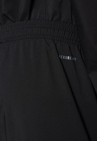 Juodas sportinis audinys turi elastingą juosmenį su susiraukšlėtu detalumu. "AEROREADY" logotipas mėlynai atspausdintas arti siūlės.