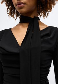 Γυναίκα με σγουρά μαλλιά που φοράει μαύρο μπλουζάκι με V λαιμόκοψη και μια μακριά μαύρη εσάρπα δεμένη γύρω από το λαιμό της, με απλό φόντο.