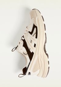 Beige athletic shoe with white mesh, dark brown suede accents, textured rubber sole, and round laces. Features a curved shape and padded collar.