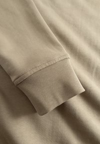 Beige bomuld langærmet skjorte detalje med fokus på cuffen, der fremhæver en glat tekstur og pæne sømme langs kanten.