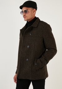 Brauner Hahnentritt-Muster Mantel mit doppelreihigen Knöpfen, breitem Kragen und Seitentaschen, getragen über einem schwarzen Rollkragenpullover.