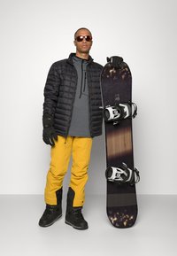 Giacca a vento nera, maglietta a maniche lunghe grigia, pantaloni da neve gialli e stivali da neve neri. Tieni in mano uno snowboard con attacchi neri e bianchi.
