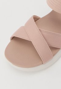 Pinkki kangassandaalit, joissa on ristikkäiset hihnat, sileä beige pohja ja valkoinen kumipohja, suunniteltu mukavuutta ja vakautta varten.