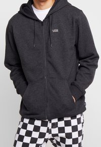 Osoba v tmavě šedé mikině na zip Vans přes bílou košili a černobílé šachovnicové kalhoty, s rukama v kapsách, před jednobarevným pozadím.