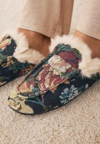Hausschuhe mit Blumenmuster, bestehend aus einem dunklen Stoffboden mit strukturierten Blumen, weichem Kunstpelzbesatz und rundem Zehendesign. Strukturierte Sohle.
