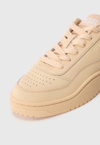 Baskets en cuir beige avec un bout perforé, des lacets plats et une semelle en caoutchouc texturé. Présente un logo en relief sur la languette et des accents cousus.