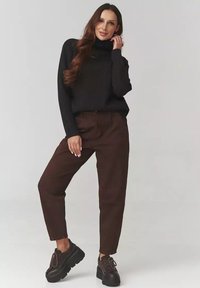 Kobieta pozująca w czarnej prążkowanej sweterze z golfem, brązowych spodniach o wysokim stanie i czarnych butach na platformie na jednolitym tle.