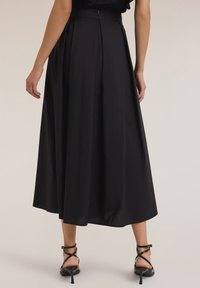 Falda negra midi con textura suave, que presenta una cintura ajustada y pliegues, combinada con sandalias de tacón negro con tiras.