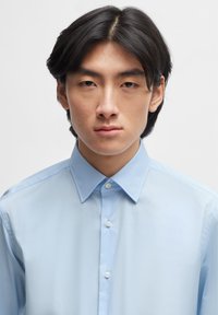 Camisa azul claro, com gola apontada, textura lisa e botões visíveis na frente, apresentando um design simples e clean.