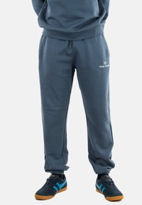 Graue Sweatpants aus einem weichen Material mit einem elastischen Bund, ausgestattet mit einem Logo auf dem linken Oberschenkel und bündchenförmigen Knöcheln.