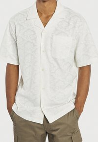 Wit shirt met korte mouwen en een gestructureerd bloemenpatroon, een borstzak en een kraag, gecombineerd met olijfgroene cargoshorts.