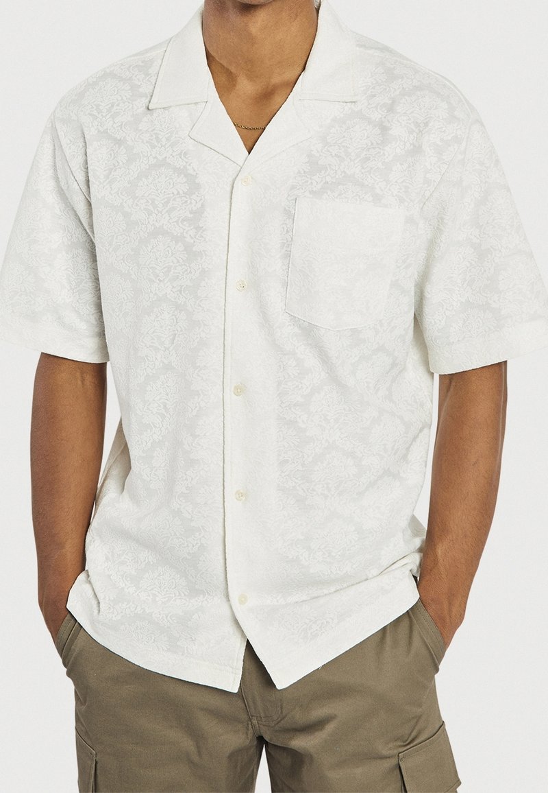 Wit shirt met korte mouwen en een gestructureerd bloemenpatroon, een borstzak en een kraag, gecombineerd met olijfgroene cargoshorts.