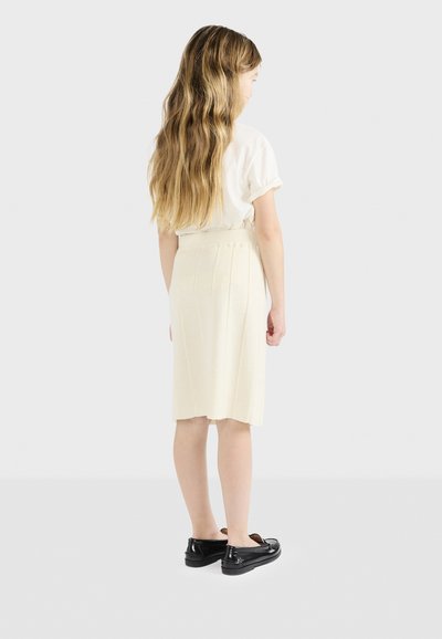 Πλεκτή κρεμ φούστα με κάθετες πιέτες, συνδυασμένη με λευκό τοπ. Μαύρα slip-on παπούτσια. Μαλλιά μακριά και κυματιστά, ορατά από το πίσω μέρος.