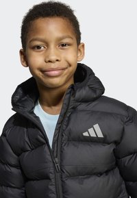 Giacca imbottita nera con collo alto, trama trapuntata, chiusura con cerniera e logo Adidas argento sul petto sinistro. Camicia azzurro chiaro sotto.