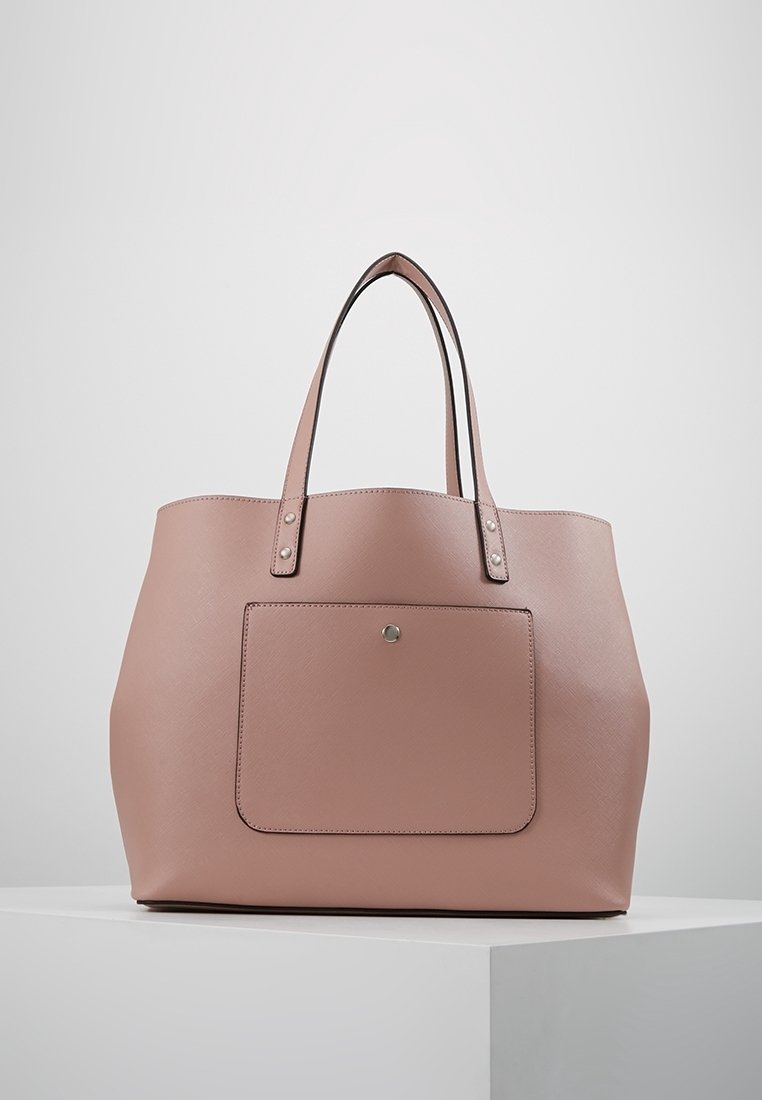 Sac cabas en cuir rose doux avec deux poignées et une poche avant sécurisée par un seul bouton-pression en métal, reposant sur une surface blanche.