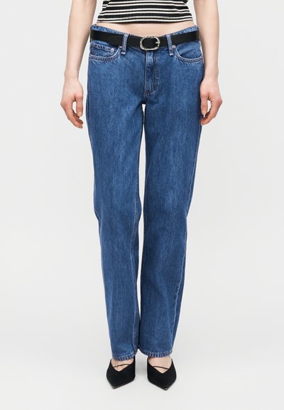 Γυναίκα που φορεί ψηλόμεσα μπλε τζιν με μαύρη ζώνη, μαύρα παπούτσια με μυτερή μύτη και ένα μαύρο-λευκό ριγωτό κοντό τοπ.