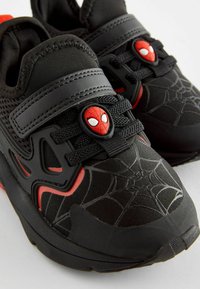 Baskets noires de sport avec des accents rouges et un motif en toile d'araignée. Les caractéristiques comprennent un emblème de Spider-Man, des lacets élastiques et une semelle rembourrée.
