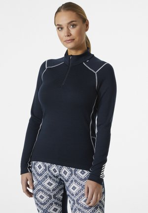 Marineblauwe pullover met lange mouwen en een kwart rits. Kenmerkt zich door witte stiksels langs de naden. Gepaard met gestreepte leggings met geometrische patronen.