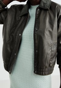 Blouson aviateur en cuir vert olive avec col et poignets côtelés, fermeture éclair à l'avant et boutons-pression. Porté sur un pull texturé vert menthe clair.