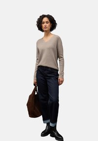 Beiger V-Ausschnitt-Pullover, dunkelblaue umgeschlagene Jeans, schwarze Stiefeletten, hält eine braune Tragetasche; glatte Textur, lässiges Design.