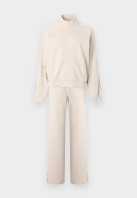 Bež trenirka z zadrgo in visokim ovratnikom, z ohlapnim krojem, dolgimi rokavi in stranskimi črtami na hlačah. Mehak tekstilni material.