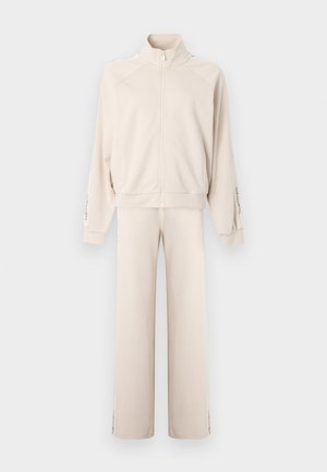 Beige hettejakke med glidelås og høy krage, med en avslappet passform, lange ermer og sidestriper på buksene. Myk stofftekstur.