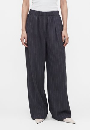 Person iført sorte vidde bukseskøjter med smalle hvide sko, stående mod en ensfarvet hvid baggrund.