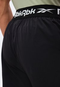 Gros plan sur un short de sport noir avec une ceinture élastique noire arborant le logo blanc de Reebok.