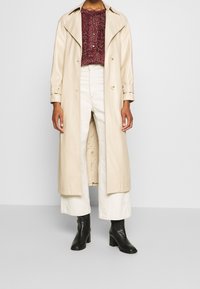Trench coat beige lungo con design doppiopetto, abbinato a un maglione in maglia testurizzata color bordeaux e pantaloni bianchi a gamba larga. Stivali neri con tacco.