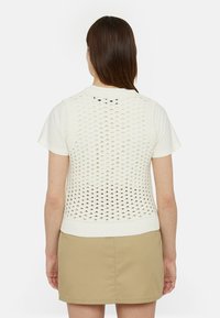 Gilet senza maniche bianco lavorato a maglia con un motivo a trama aperta e orlo a coste, abbinato a una maglietta leggera e una gonna beige.