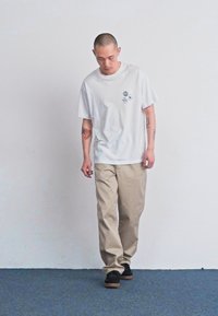 White cotton t-shirt with three small graphics on the chest, paired with light beige pants and black sneakers, standing on blue carpet.