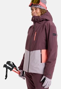Purple and light gray ski jacket with orange accents, zippered pockets, and adjustable cuffs, paired with pink ski goggles and poles.