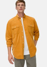 Senfgelbes Hemd mit Knöpfen aus strukturiertem Stoff, das zwei Brusttaschen und rollbare Ärmel hat. Getragen über einem weißen T-Shirt.