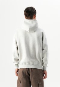 Osoba z krótkimi ciemnymi włosami, ubrana w jasnoszary hoodie i brązowe spodnie cargo, stojąca tyłem na jednolitym białym tle.