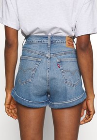 Shorts en denim bleu clair avec des poignets retroussés, dotés de deux poches arrière et d'un patch en cuir brun sur la taille.