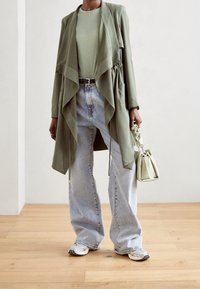 Olivgrüne lange Jacke mit gerafftem Vorderteil, kombiniert mit einem hellgrünen Hemd, locker sitzenden hellblauen Jeans und einer kleinen hellgrünen Handtasche.