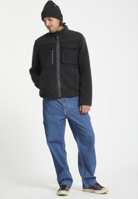 Veste en polaire noire avec fermeture éclair sur le devant, deux poches poitrine et panneaux en tissu noir ; assortie à un jean bleu ample et des baskets sombres.