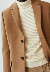 Lys brun blazer med reverskrave og to sorte knapper, lagd oven på en struktureret cremefarvet turtle-neck sweater med ribbet mønster.