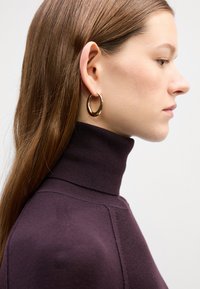 Boucle d'oreille en or avec une finition polie, portée avec un pull à col montant prune foncé ; texture lisse et forme légèrement courbée.