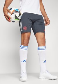 Grå idrottsshorts med röd Manchester United-logga, kombinerat med vita strumpor med blå ränder och vita fotbollsskor, håller en fotboll.