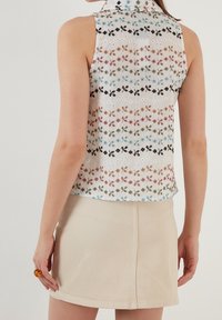 Bez rękawów bluzka z wielokolorową haftowaną florystyką na białym materiale; posiada kołnierzyk i lekko luźny krój, zestawiona z beżową spódnicą.