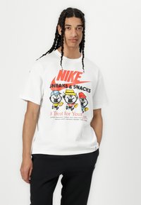 Hvit bomull t-skjorte med grafisk trykk av tre tegneseriefigurer med mat og en dristig rød "NIKE"-logo. Korte ermer, avslappet passform.