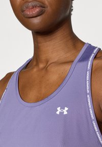 Μωβ αθλητική φανέλα χωρίς μανίκια με ομαλή υφή, που διαθέτει λευκή σήμανση "Under Armour" στα λουριά και λευκό λογότυπο στο στήθος.