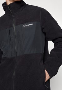 Giacca in pile nera con chiusura a zip, colletto alto e pannelli delle spalle neri a contrasto. Presenta un logo sul petto e tasche laterali.